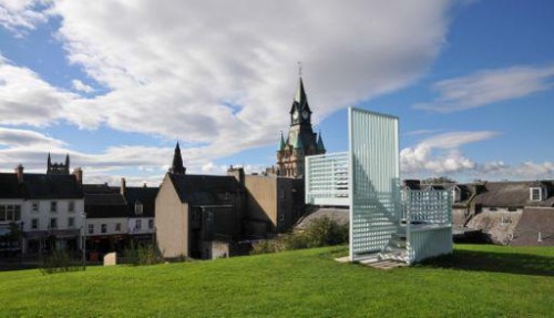 External photo of abstract large light-blue structure set on top of hill overlooking town with church steeple prominent