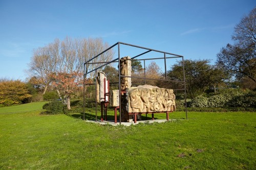 External photo of large abstract structure comprising stone walls within metal framework set in grassy parkland with trees in background